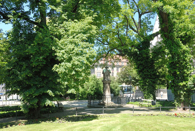 The Old Bach Monument stands under trees in a green area in Leipzig. In the foreground is lawn, in the background are houses, it is quite small in the picture and difficult to recognize.