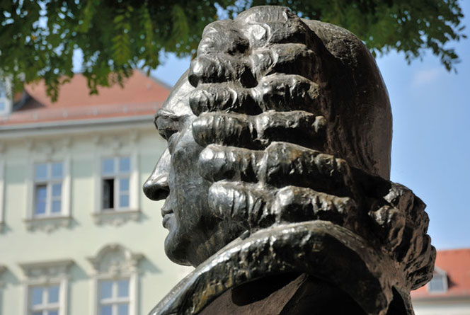 The Bach monument in Weimar. Bach is photographed at a slight angle from the back. In the background is a historic house, above the foliage of a tree and a cloudless sky.