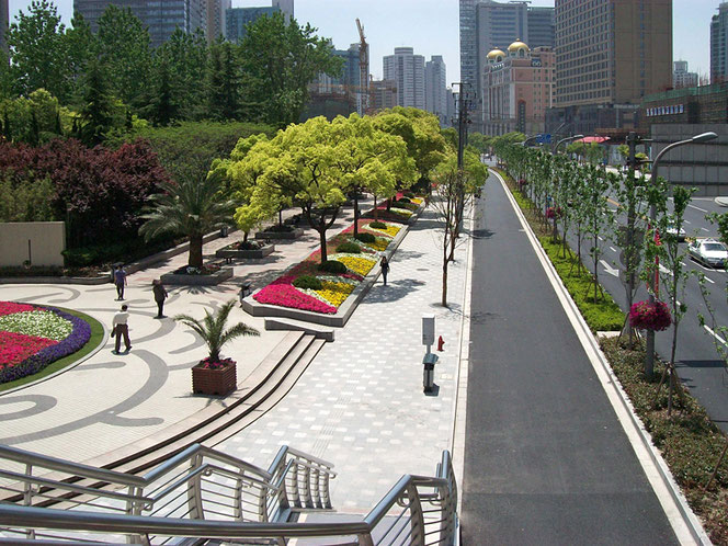 A park in Shanghai in China is in the left half of the picture, on the right is a street. Everything is very well maintained and beautifully planted. You can hardly see any people. There are skyscrapers in the background.