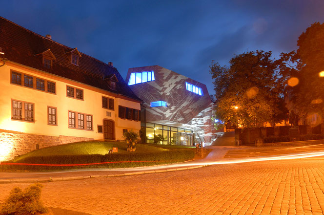 The Bach ensemble on the Frauenplan in Eisenach after dusk. Everything is brightly lit: the Bach House on the left and the Bach Museum. Only the monument is barely visible in this photo. The sky is a deep dark blue.