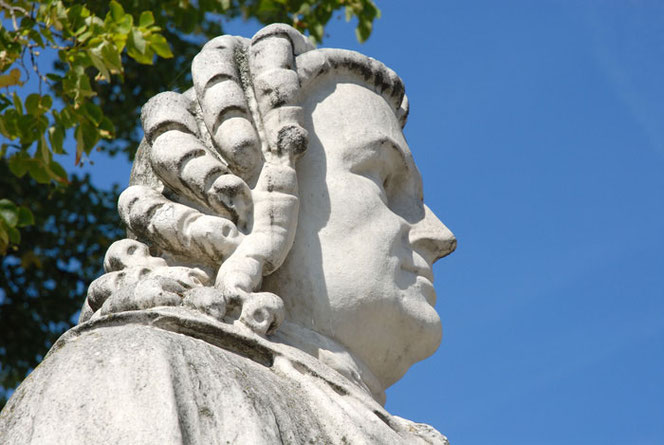 The head to the chin of the Bach monument in Köthen. It is white, and the shadows make it even more three-dimensional. On the right is blue sky, on the left the spring foliage of a tree.