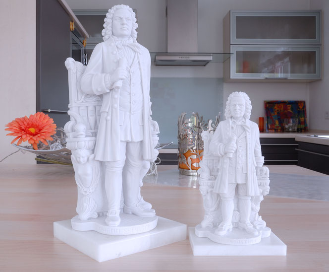 Two Bach statues of different sizes stand on an island in a kitchen. Both represent the New Bach Monument in Leipzig. Both are made of white Gypsum. In the background is a bowl with a pink decorative flower, on the right you can see the kitchen.
