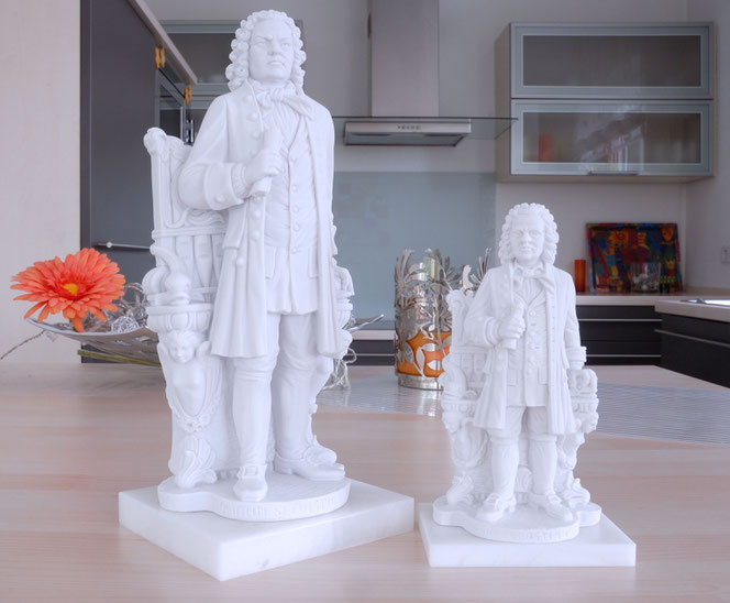 Two Bach statues of different sizes stand on an island in a kitchen. Both represent the New Bach Monument in Leipzig. Both are made of white Gypsum. In the background is a bowl with a pink decorative flower, on the right you can see the kitchen.