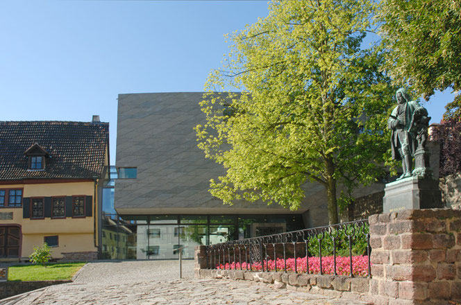 The Bach ensemble in Eisenach on the Frauenplan: On the left is a third of the yellow Bach House, in the middle is the modern, gray Bach Museum, on the right is the Bach monument. Between the museum and the monument is a tree in spring foliage.