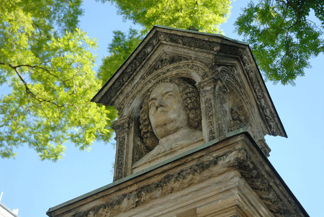 The view is upwards, diagonally towards the portrait of Bach at the Old Bach Monument. There is a cloudless sky in the background and trees in spring foliage.