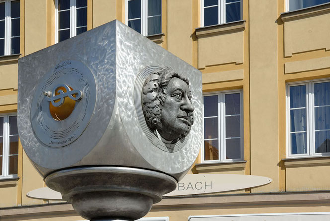 The Bach monument in Ansbach, Bavaria, Germany. Silver, a cube with a semi-circular lower half. You look at two sides, one is the portrait. Behind it is a yellow, historical building and you can read BACH.