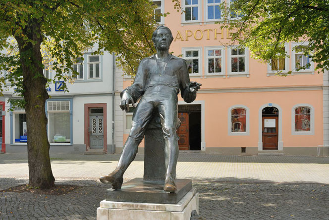 The monument dedicated to Bach in Arnstadt. It stands on a paved area in front of a salmon-colored house. Bach lounges standing on his monument pedestal and looks past the viewer.