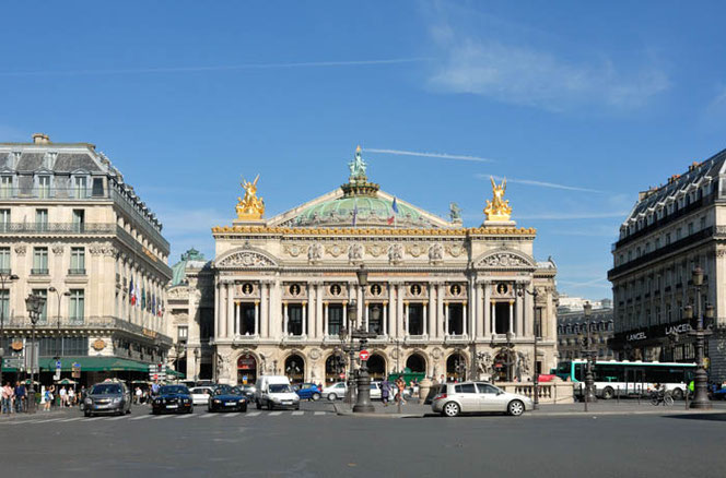 A view onto the Paris Opera in France in the sunshine. In front, traffic flows on the forecourt, to the right and left are historic residential buildings. The sky is blue.