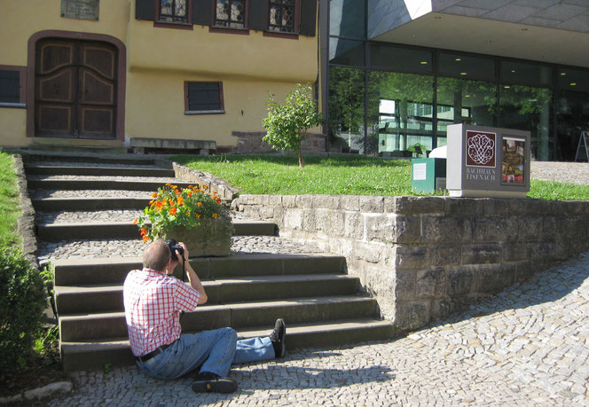Peter Bach Jr. sits on the ground in front of the Bach House, the Bach Museum and a large information box and photographs the motif from an exciting perspective.