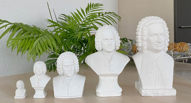 Five Bach gypsum busts stand on a beige furniture surface. There are five different sizes. The smallest bust is on the left. In the background on the left is a palm tree, on the right a bowl with decoration and a cupboard door.