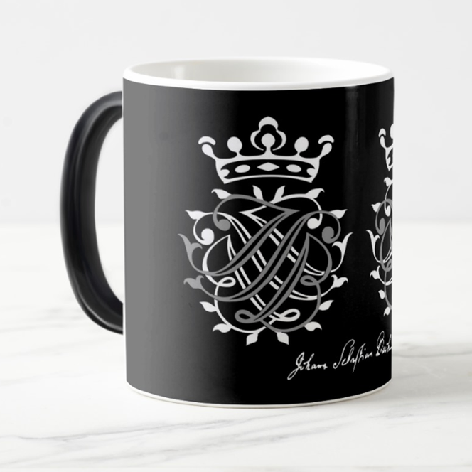 A large black cup with a handle stands on a white background. The decoration is the Bach Seal in white. On the right edge of the cup, you can see that there is a second seal on the other side. At the bottom is the signature of Johann Sebastian Bach