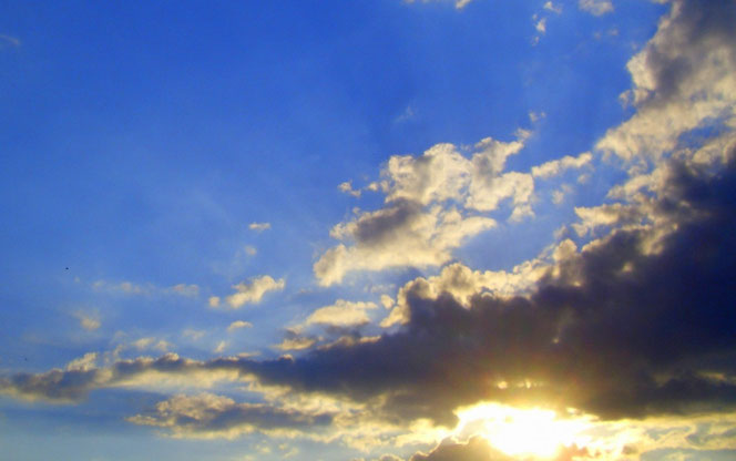 Sonne bricht durch dunkle Wolken am blauen Himmel.