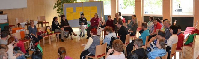 Gruppe von Erwachsenen und Kindern sitzt in einem Stuhlkreis in einem hellen Raum; die Teilnehmenden wirken in ein gemeinsames Gespräch oder eine moderierte Gruppenaktivität eingebunden.