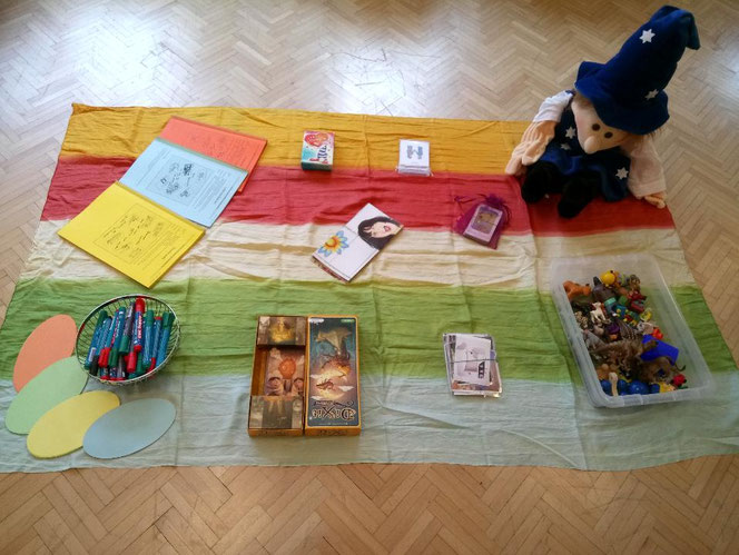 Materialien für eine pädagogische Einheit auf bunter Decke ausgebreitet, darunter Karten, Stifte, Hefte, Spielfiguren und eine Stoffpuppe.