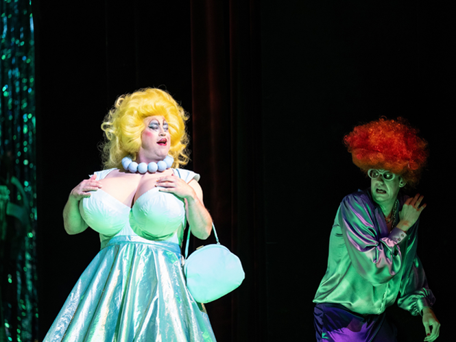 Zwei Schauspieler in Kostümen auf der Bühne: Einer mit gelber Perücke und glitzerndem Kleid, der andere mit roter Lockenperücke und grünem Make-up.