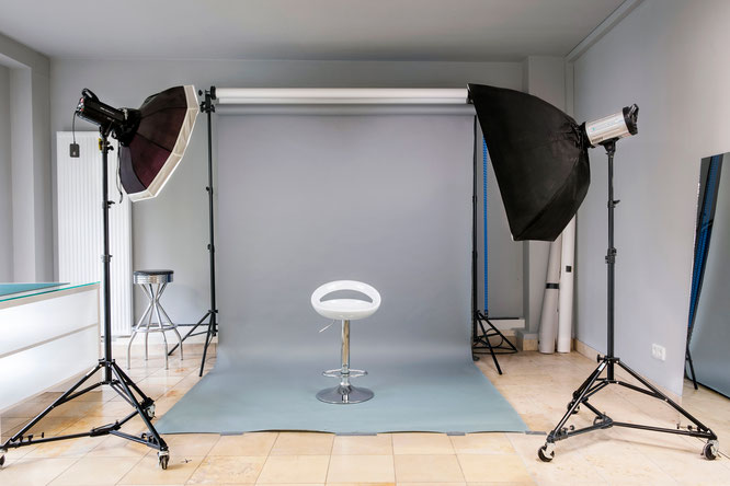 Das Bild zeigt ein professionell eingerichtetes Fotostudio mit diversen Beleuchtungsausrüstungen wie Softboxen und einem Beauty Dish, gerichtet auf einen Hintergrund und einen weißen Hocker in der Mitte, ideal für Porträtfotografie oder Produktaufnahmen.