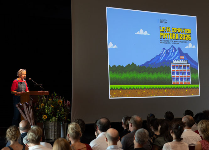 Rektorin Annette Studer im Super-Mario-Kostüm bei der Eröffnungsrede der Maturafeier 2026 der Kantonsschule Reussbühl vor einer Leinwand mit dem Motto «Level Completed».