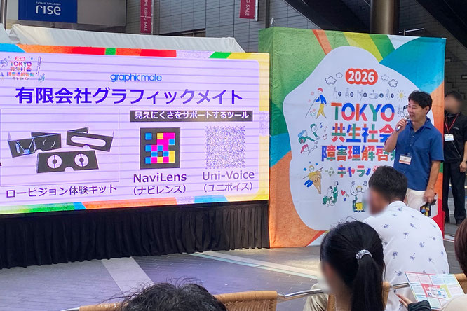 イベント会場のステージの様子。ステージ上のパネルには「有限会社グラフィックメイト」の社名や「ロービジョン体験キット」、「NaviLens（ナビレンス）」、「Uni-Voice（ユニボイス）」の名称とコードが記載されている。ステージ右側には男性がマイクを持って話しています。男性の後ろには「TOKYO共生社会 障害理解啓発キャラバン」と書かれたカラフルな看板が設置され、手前には観客の姿も見受けられます。