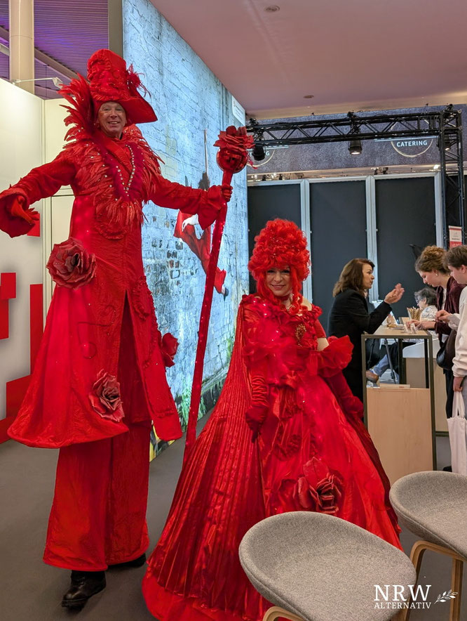 ZWei ganz in rot gekleidete Stelzenläufer an einem Messestand.