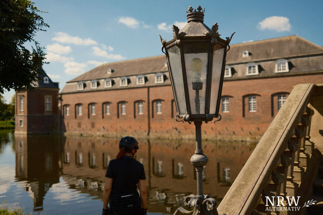 Frau mit Fahrradhelm, die ins Wasser eines Schlossgrabens schaut, in dem sich das Schloss spiegelt.