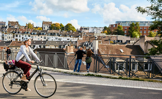 Eine Frau sitzt auf einem Fahrrad und lächelt. Im Hintergrund sind Hausdächer zu sehen. 