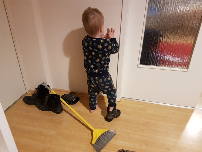 Zweijähriges Kleinkind mit angezogenem Winterstiefel und ungünstig platziertem Besen in Wohnung.