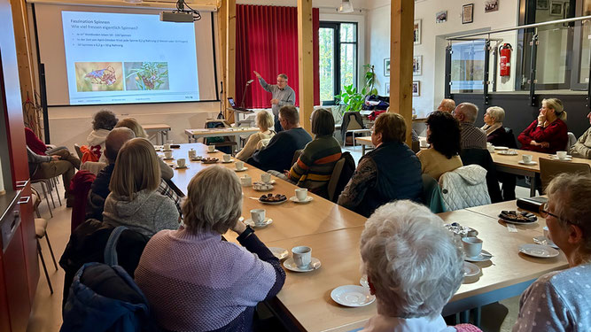 Der Referent erläutert einzelne Punkte der Präsentation vor einem gemischten Publikum. Der Raum ist hell, auf den Tischen stehen Kaffee und Gebäck.