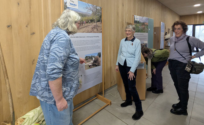 Zwei Frauen schauen einer anderen Frau zu, wie sie mit der Hand auf Banner einer Ausstellung zeigt. Die Frau scheint den anderen Frauen, die lächeln, etwas zu erklären. 