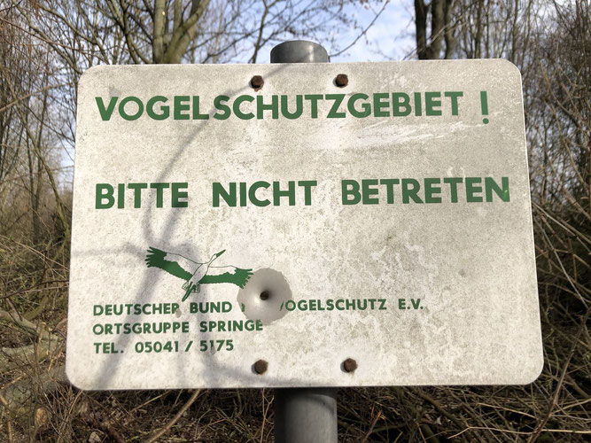 Altes Warnschild in weiß mit grüner Schrift, in der Mitte ist ein großes Einschussloch