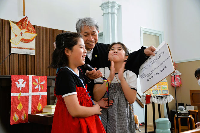 2026年6月14日(日)こども祝福礼拝にて 創世記1章31節のお話をする森牧師と前方に招かれた双子ちゃん