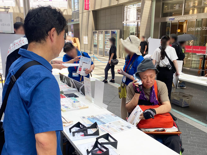 屋外の通路に設置されたテーブルに、ロービジョン体験キットが置かれている。手前には当社スタッフの男性が立ち、テーブルの向こう側には車椅子ユーザーの男性。当社のニュースレターに掲載された音声コードUni-Voiceをスマホで読み取り、スマホを耳に当てて音声を聞いている。
