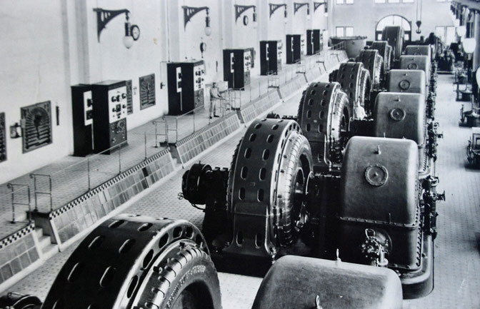Blick in den damaligen Maschinenraum mit sieben horizontalachsigen Pelton-Turbinen. Das Bild zeigt jeweils das Turbinen(gehäuse) rechts, den Generator links.