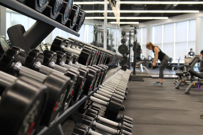 Aufgeräumter Freihantelbereich in einem Fitnessstudio. Im Hintergrund sieht man eine Frau, die trainiert.