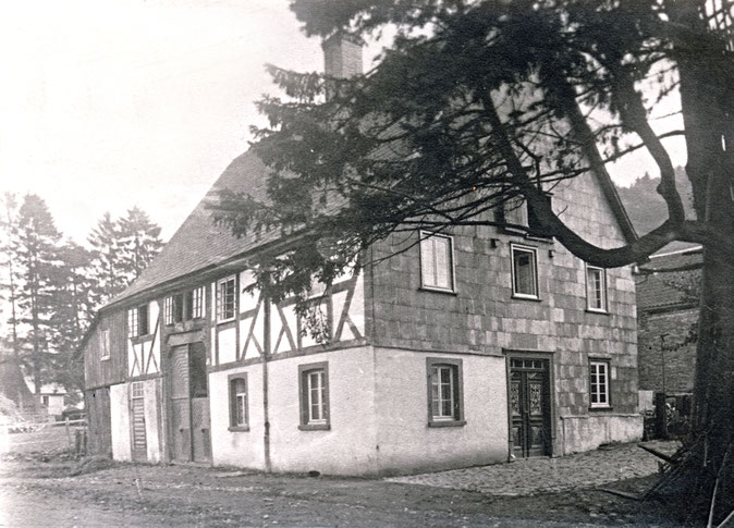 So sah „Schmies Haus“ vor dem Umbau 1910 aus: Der Hauseingang auf der nach Südwesten ausgerichteten Giebelseite, ein großes Deelentor und der Eingang zum Stall an der Straßenseite. Im Hintergrund erkennt man die bis zur Straße reichende Reihe hohe Fichte.