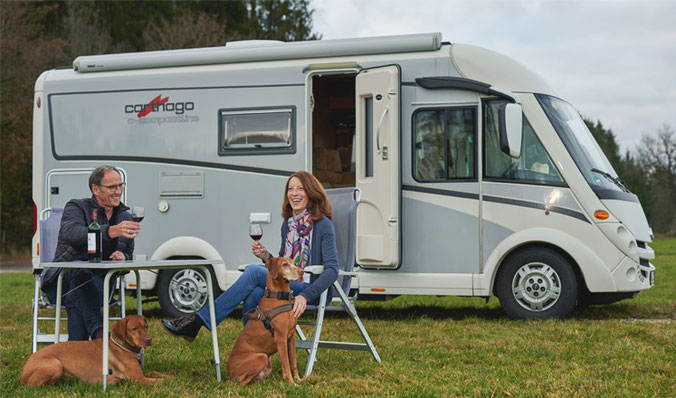 Ein älteres Ehepaar mit seinen zwei Hunden vor ihrem Caravan bei einem Glas Wein. Sie genießen aufgrund guter Altersvorsorge sorgenfrei ihren Ruhestand