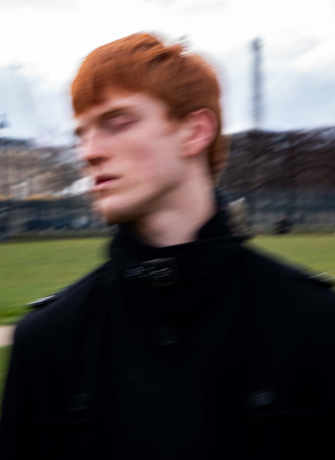 Color fine art portrait of a professional male model with red hair, captured in motion blur, wearing a dark coat outdoors against a muted landscape