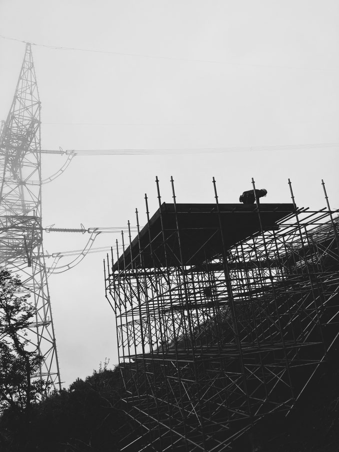 霧がかった空の下、高所に組まれた複雑な足場の上で作業をする足場職人のシルエット