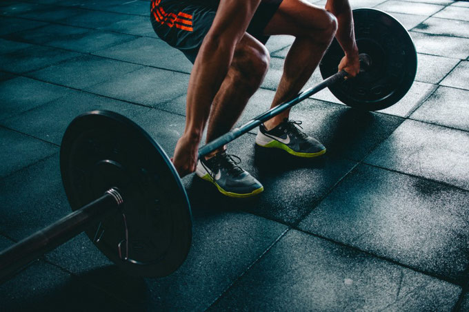 Man sieht den Unterkörper eines Athleten in Sportkleidung, der zum Kreuzheben einer Langhantelstange mit Gewichten ansetzt.