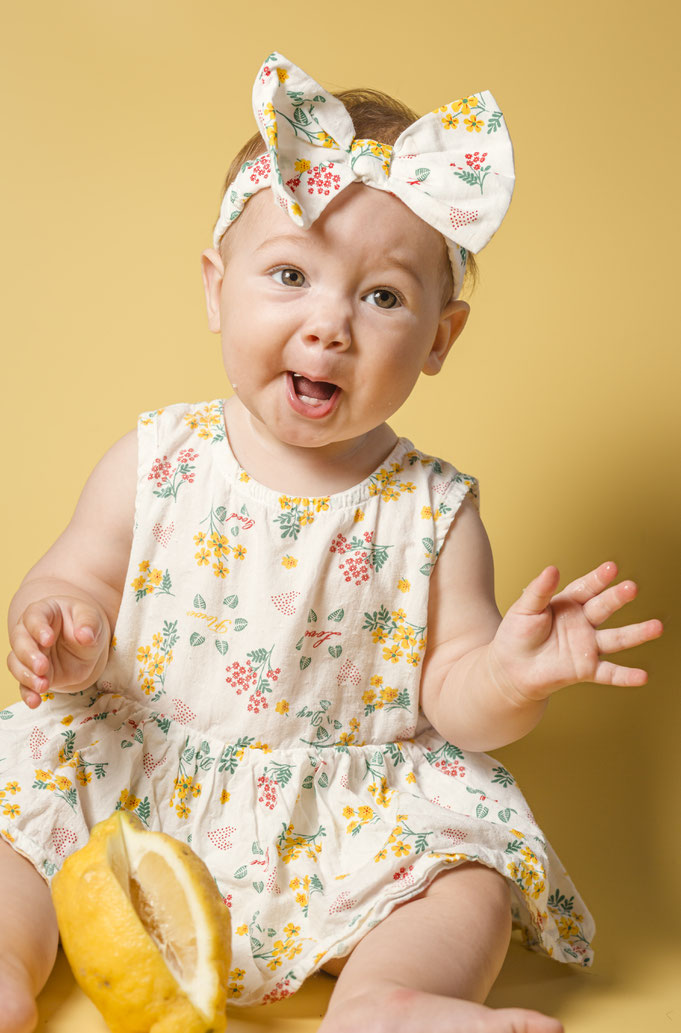 Baby in weißem Sommerkleid mit Bunten Blumen drauf. Sie sitzt vor gelben Hintergrund mit einer Zitrone in der Hand und macht ein lustiges Gesicht. Foto von Ralf Weichselbaumer