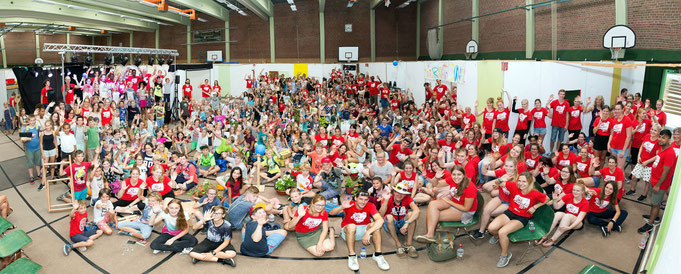 Panorama-Foto der Abschlussfeier, Mini-Regensburg 2017; viele Kinder, Jugendliche sowie Mitarbeiterinnen und Mitarbeiter sitzen auf Stühlen oder am Boden in der großen Turnhalle undwinken in die Kamera