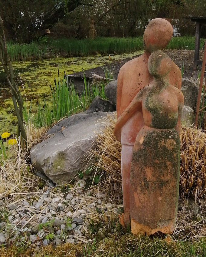 Zwei abstrahierte Steinskulpturen von einem Paar am Rand eines Teichs mit Schilf und Wasserpflanzen.