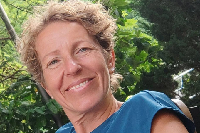 Porträt der lächelnden Christine Harnik mit kurzen, lockigen Haaren im Freien vor grünem Laub.