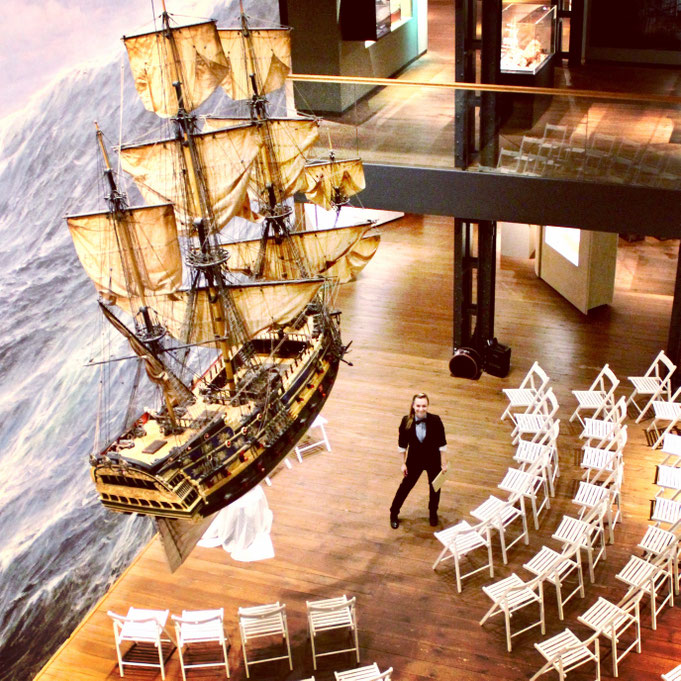 Constanze Marienfeld strahlt in die Kamera im Maritim Museum, über ihr hängt ein Segelschiff, neben ihr sind schon die Stühle für die Trauung aufgebaut.