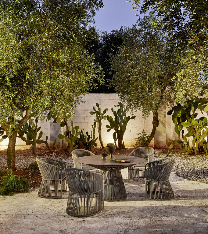 Giardino arredato con tavolo outdoor, sedie intrecciate e vegetazione mediterranea.