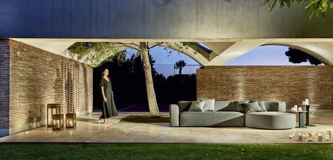 Divano outdoor sotto porticato con tavolino e parete in mattoni, soluzione relax per spazi esterni coperti.