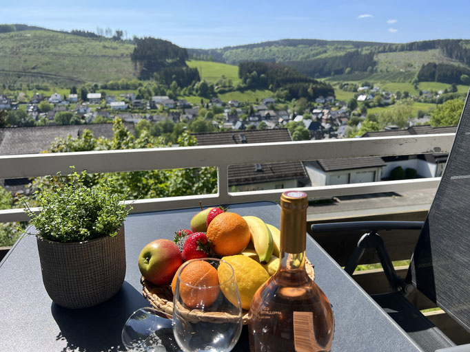 Ein Balkon mit herrlichem Blick auf die grünen Hügel und das Dorf Niedersfeld im Tal. Auf dem Tisch stehen eine Obstschale mit frischen Früchten, eine Flasche Roséwein, zwei Gläser und eine Topfpflanze – perfekt für eine entspannte Auszeit in der Sonne.