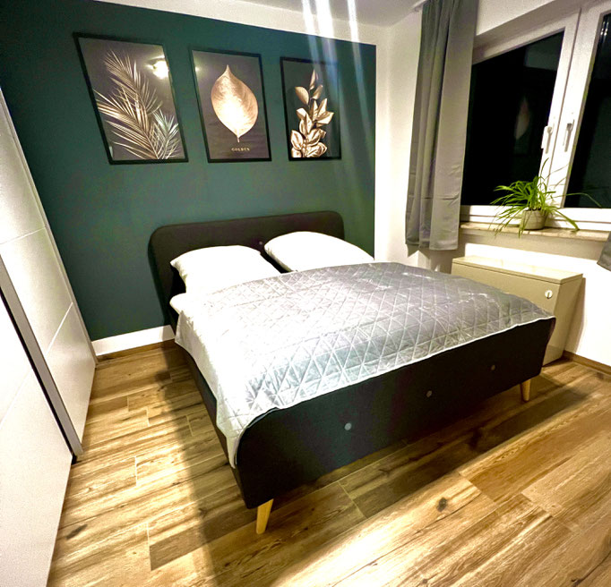 Modernes Schlafzimmer mit Doppelbett, grüner Akzentwand und stilvollen Pflanzenbildern in unserer FeWo in Winterberg. Holzfußboden, große Fenster mit Vorhängen und eine Zimmerpflanze sorgen für eine gemütliche Atmosphäre.