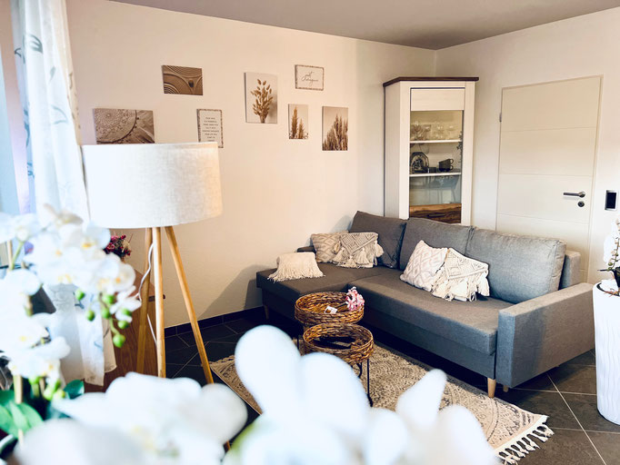 Stilvoll eingerichtetes Wohnzimmer mit grauer Couch, heller Stehlampe und dekorativen Wandbildern. Warme Farben, gemütliche Kissen und liebevolle Details schaffen eine einladende Atmosphäre in unserer Ferienwohnung in Winterberg-Niedersfeld.