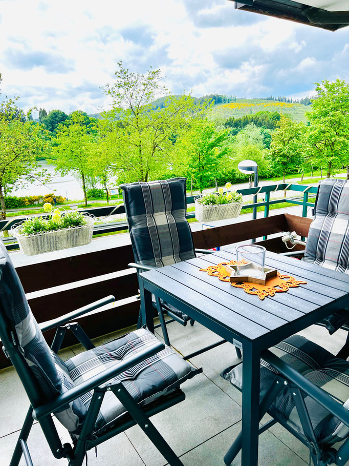 Gemütlich möblierter Balkon mit Blick ins Grüne und auf den Hillebachsee in Niedersfeld. Bequeme Hochlehner-Stühle, ein gedeckter Tisch und Blumenkästen sorgen für eine einladende Atmosphäre – ideal zum Entspannen im Sauerland.