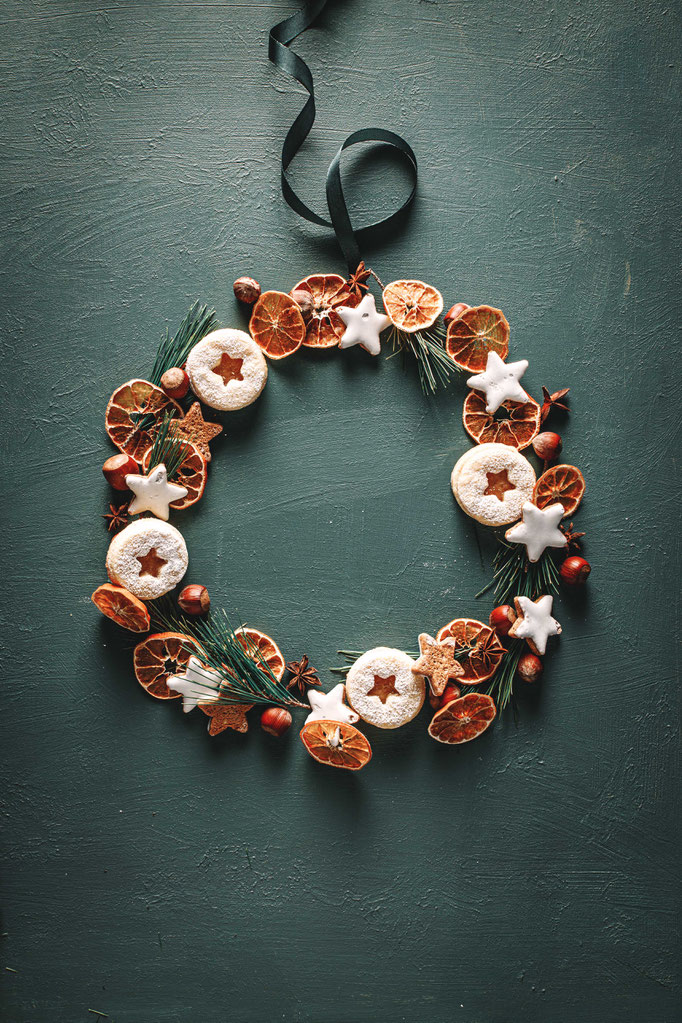 Selbstgemachter Adventskranz aus Weihnachtsplätzchen, getrockneten Orangenscheiben, Nüssen und Gewürzen vor einem grünen Hintergrund.